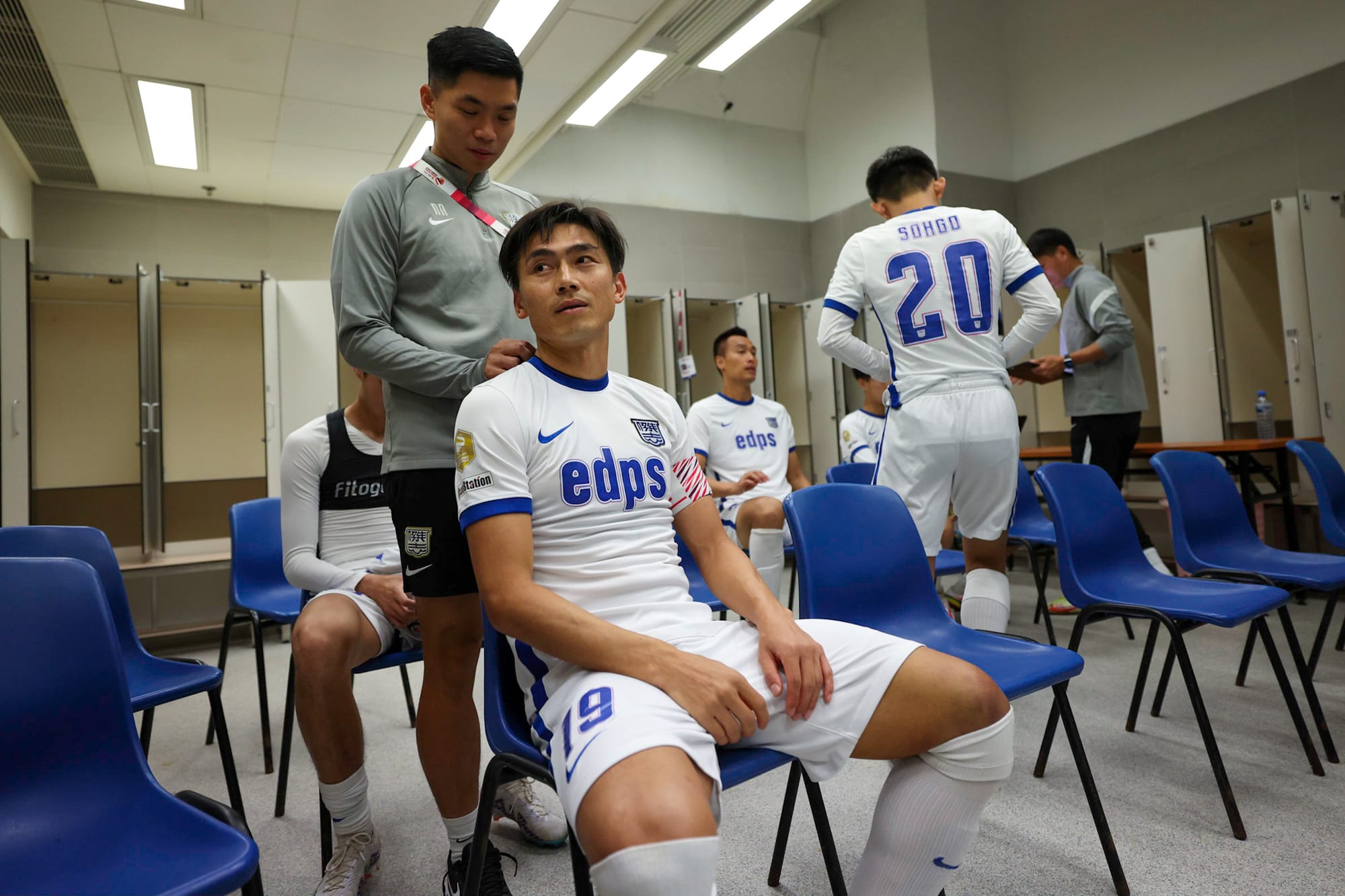 Norris Ng in a grey Kitchee training top standing behind a seated Kitchee FC player in a white edps shirt, attending to the player's shoulder in the dressing room before a match.