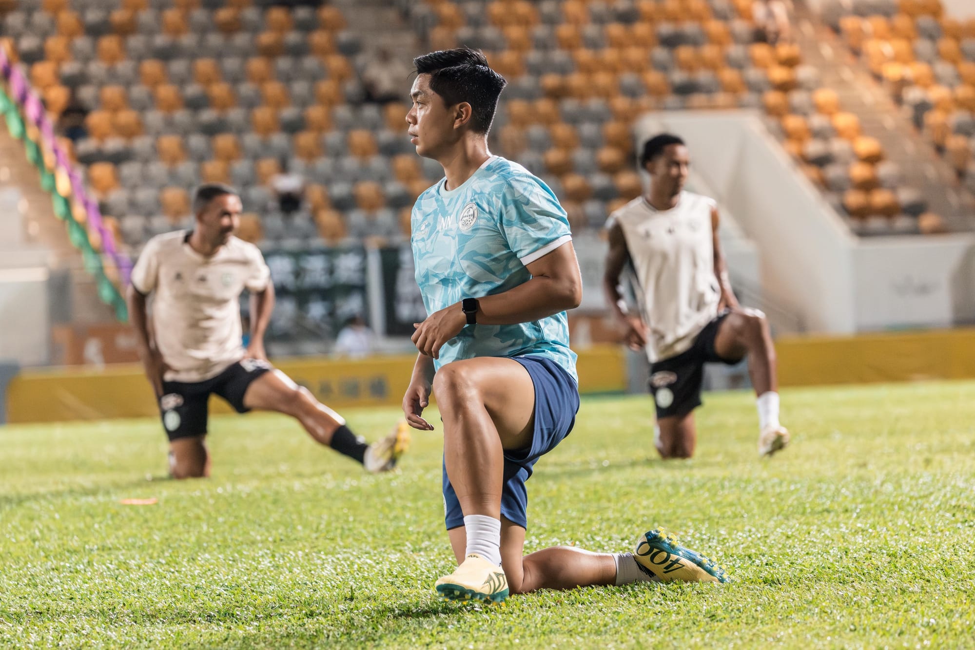 Norris Ng in a light blue Tai Po FC training shirt and blue shorts holding a deep lunge stretch on a stadium pitch, two players visible in the background doing the same drill.
