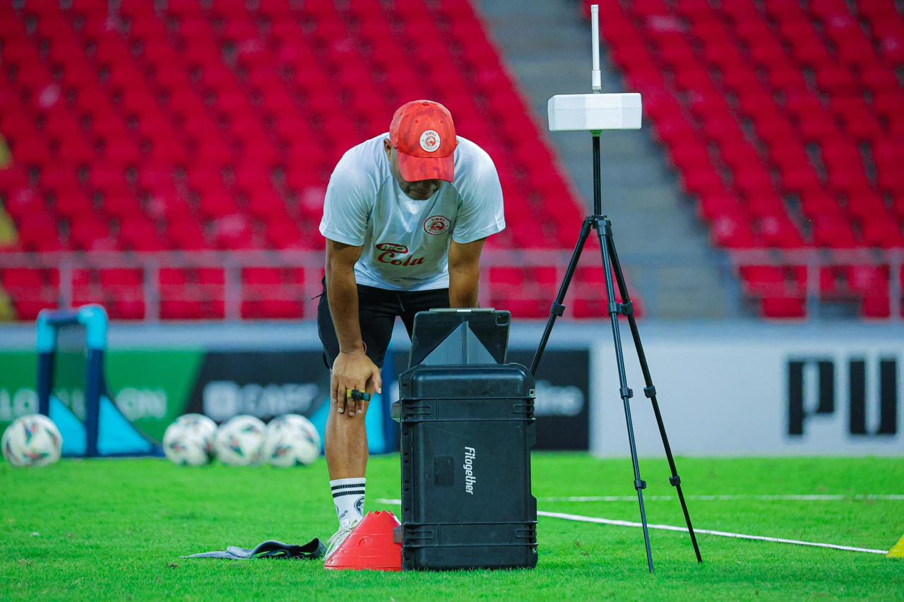 Riedoh Berdien in a red Simba cap and white club polo setting up a Fitogether GPS tripod and equipment case on an empty pitch with rows of red stadium seats behind him.