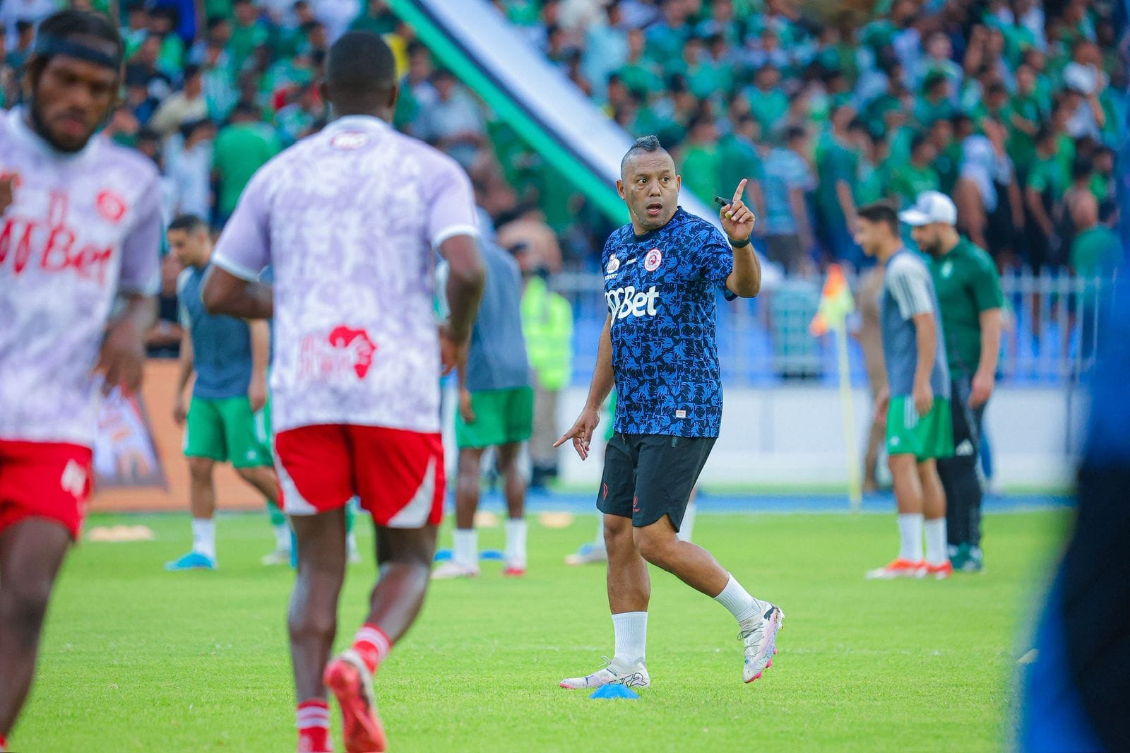 Riedoh Berdien in a blue patterned Simba Sports Club training shirt directing players during a pre-match warm-up before a packed stadium of green-clad supporters.