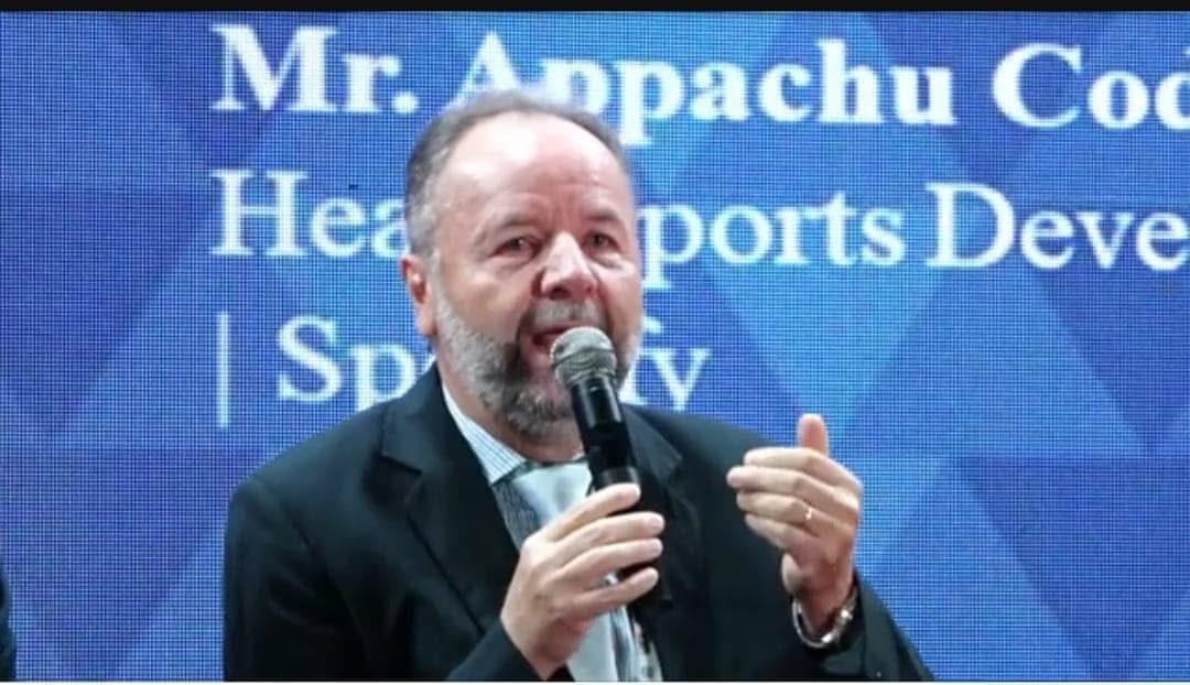 Luiz Greco speaking into a microphone at a sports development conference, blue stage backdrop visible behind him.