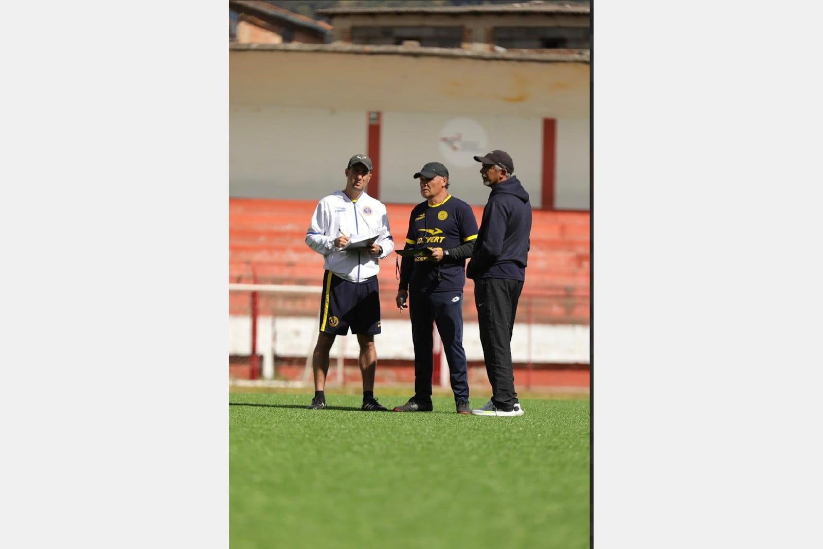 Marco Gracia in a white FC Cajamarca jacket holding a notebook, in conversation with two senior staff members on a stadium pitch in Peru.