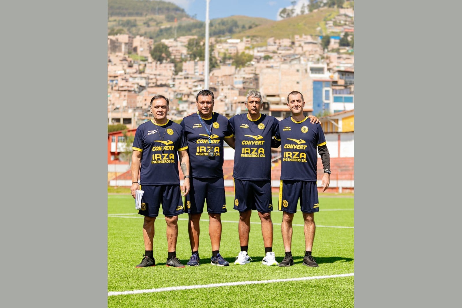 Four FC Cajamarca coaching staff members in matching navy training kit standing on a grass pitch with the city of Cajamarca, Peru rising in the background, Marco Gracia on the far right.