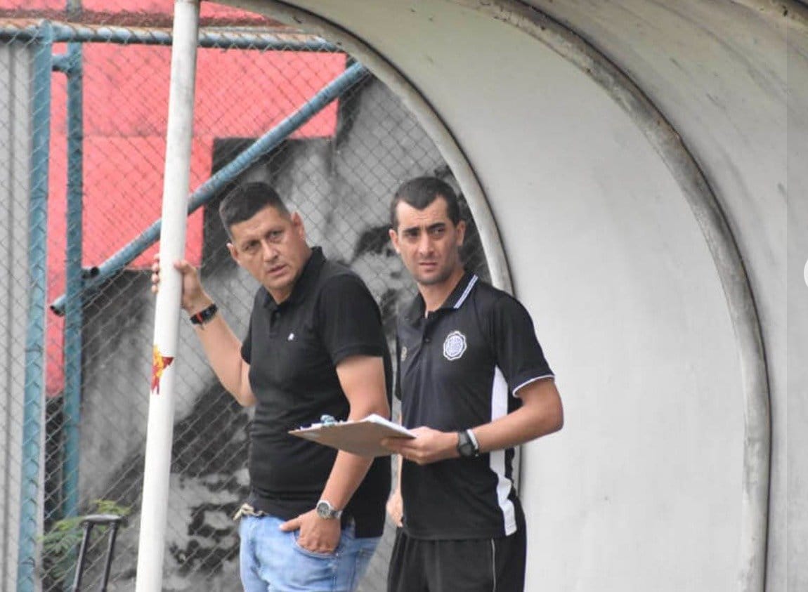 Marco Gracia in a black coaching polo standing in the dugout next to an older staff member who holds a clipboard, with a metal fence and goalpost in the foreground.