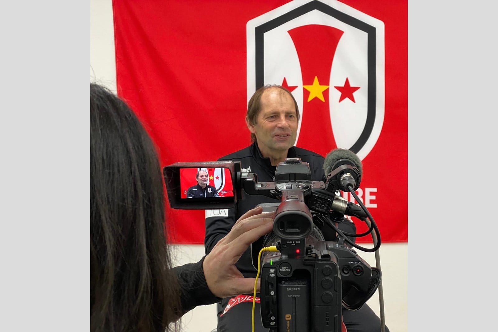 Gert Engels seated in front of a red club banner being filmed by a Sony broadcast camera, with the camera operator's hair visible in the foreground and the camera's small viewfinder showing his image.