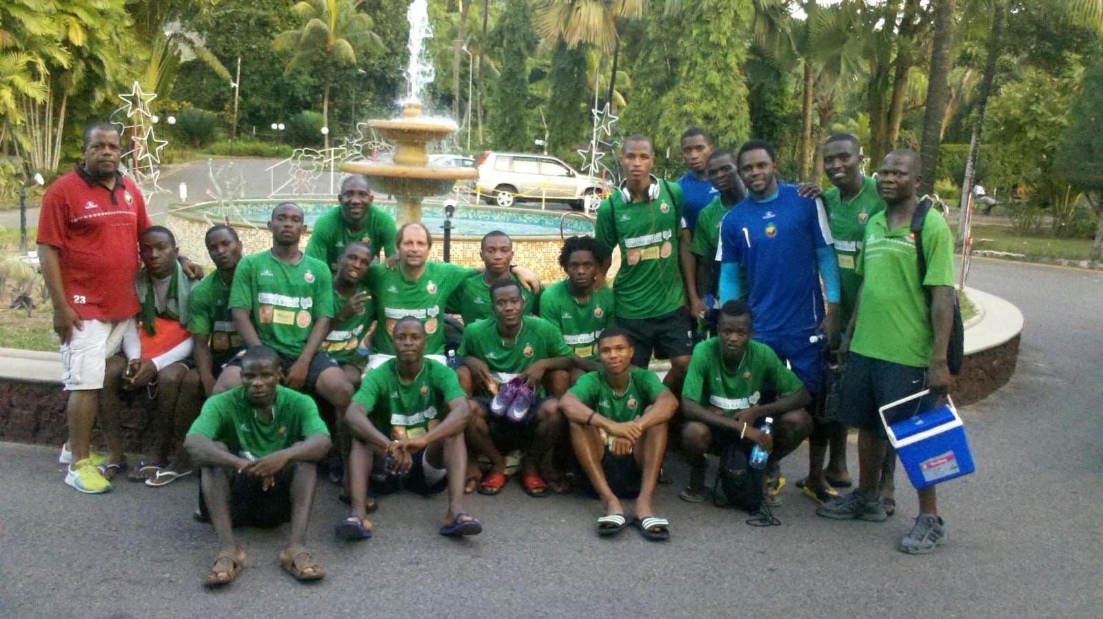 Gert Engels, in the centre of the group, posing with the Mozambique national football team squad in green training kit outside a hotel with a fountain and palm trees in the background.