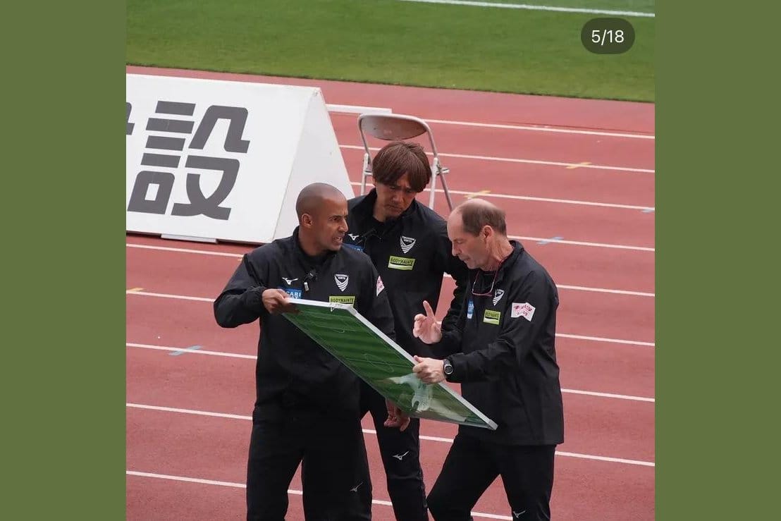 Gert Engels in a black club coaching polo holding a green tactical board, in animated conversation with two assistant coaches on the stadium track during a J.League match.