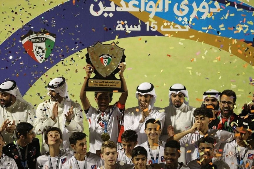 A Kuwait FA U-15 championship celebration scene with the captain raising the trophy, confetti falling, players in the foreground smiling, and Kuwaiti officials in white traditional dress applauding behind them.