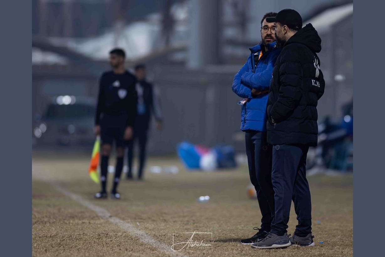 Rui Gomes in a blue puffer jacket standing on the pitch line in conversation with a colleague in a black jacket, with a referee visible in the background and the floodlit pitch fading into evening light.