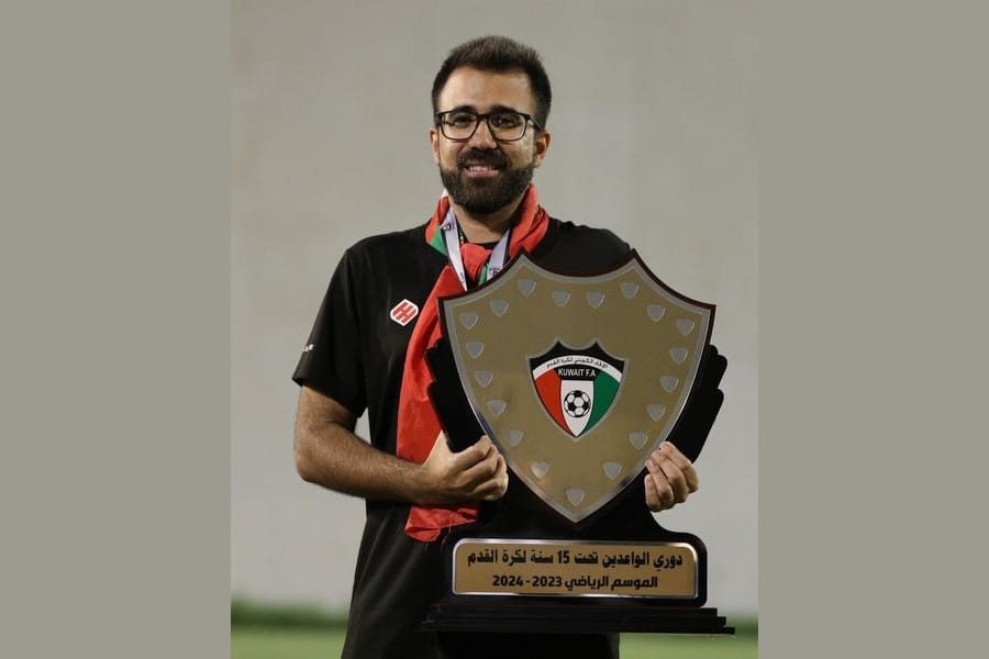 Rui Gomes in a black coaching polo, with a Kuwait Football Association lanyard around his neck, holding the Kuwait FA U-15 league championship shield in front of him on the pitch after the title-deciding match.
