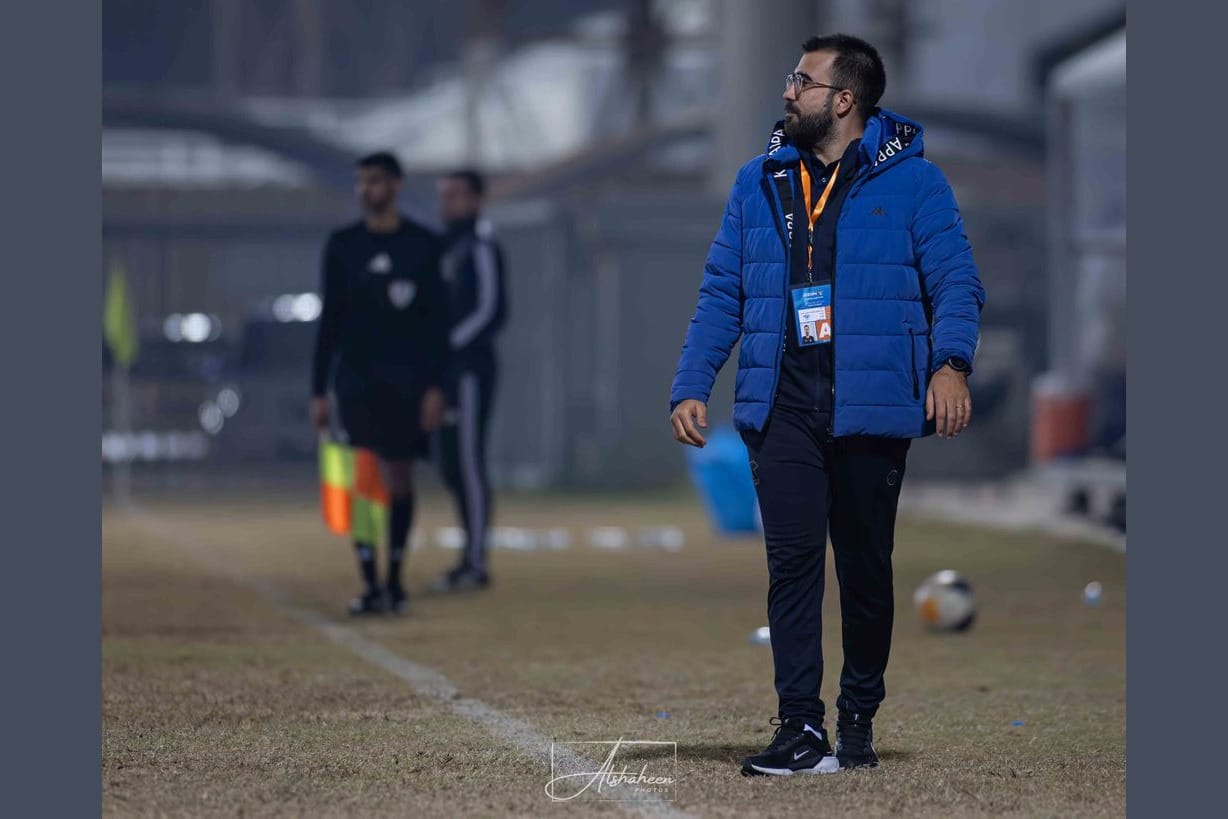Rui Gomes in a blue puffer jacket walking along the touchline of a floodlit pitch in cold weather, with a referee in the background.