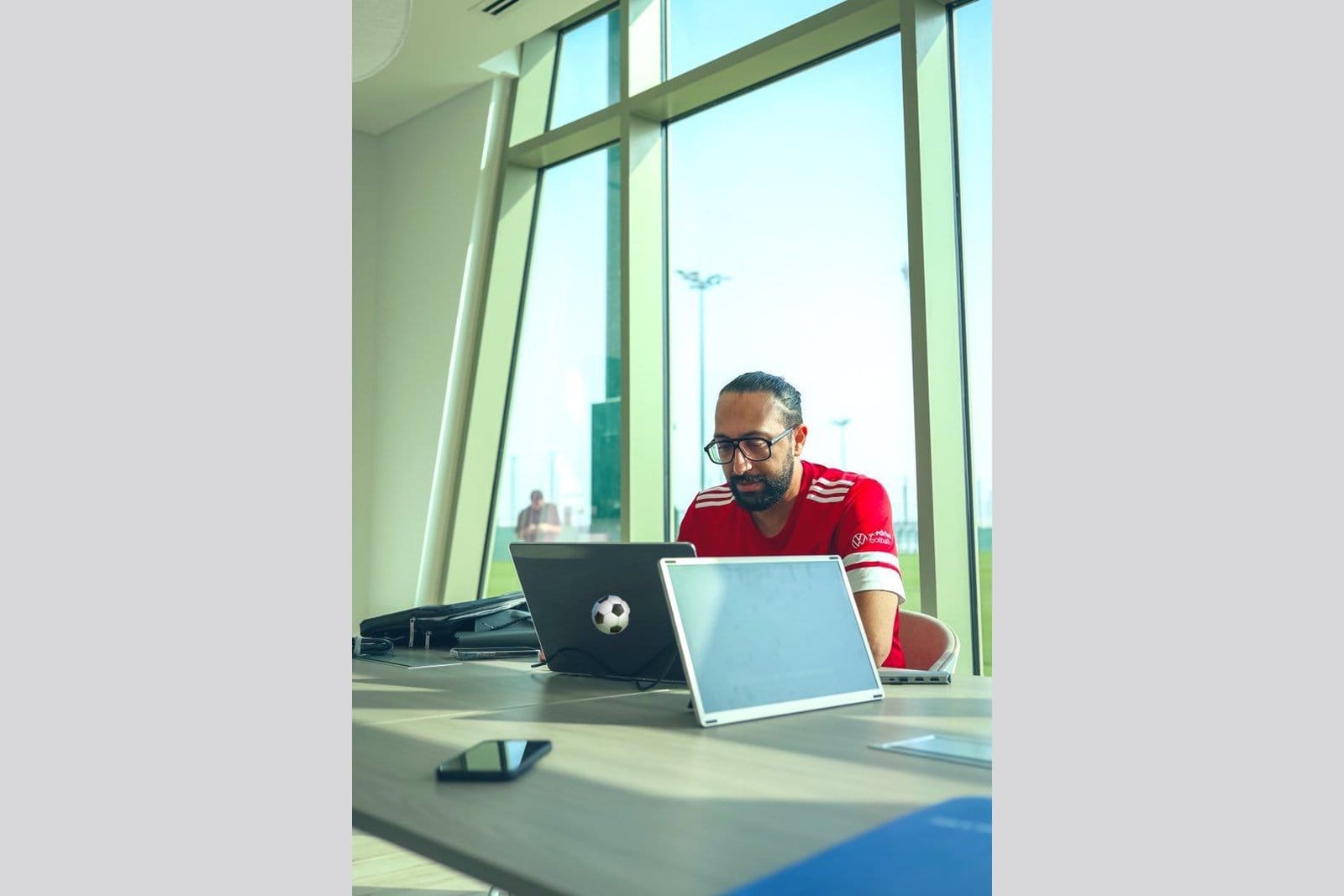 Sameh Abdelghany in a red Gulf United FC training shirt seated at a desk with two laptops, a phone, and a small football, working with stadium lights visible through the window behind him.
