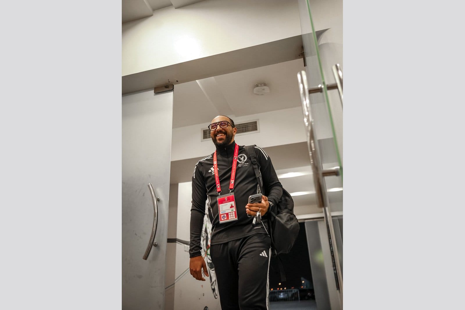 Sameh Abdelghany in an adidas Gulf United FC tracksuit and red UAE FA accreditation lanyard, carrying a backpack and smiling as he walks through a stadium entrance area.