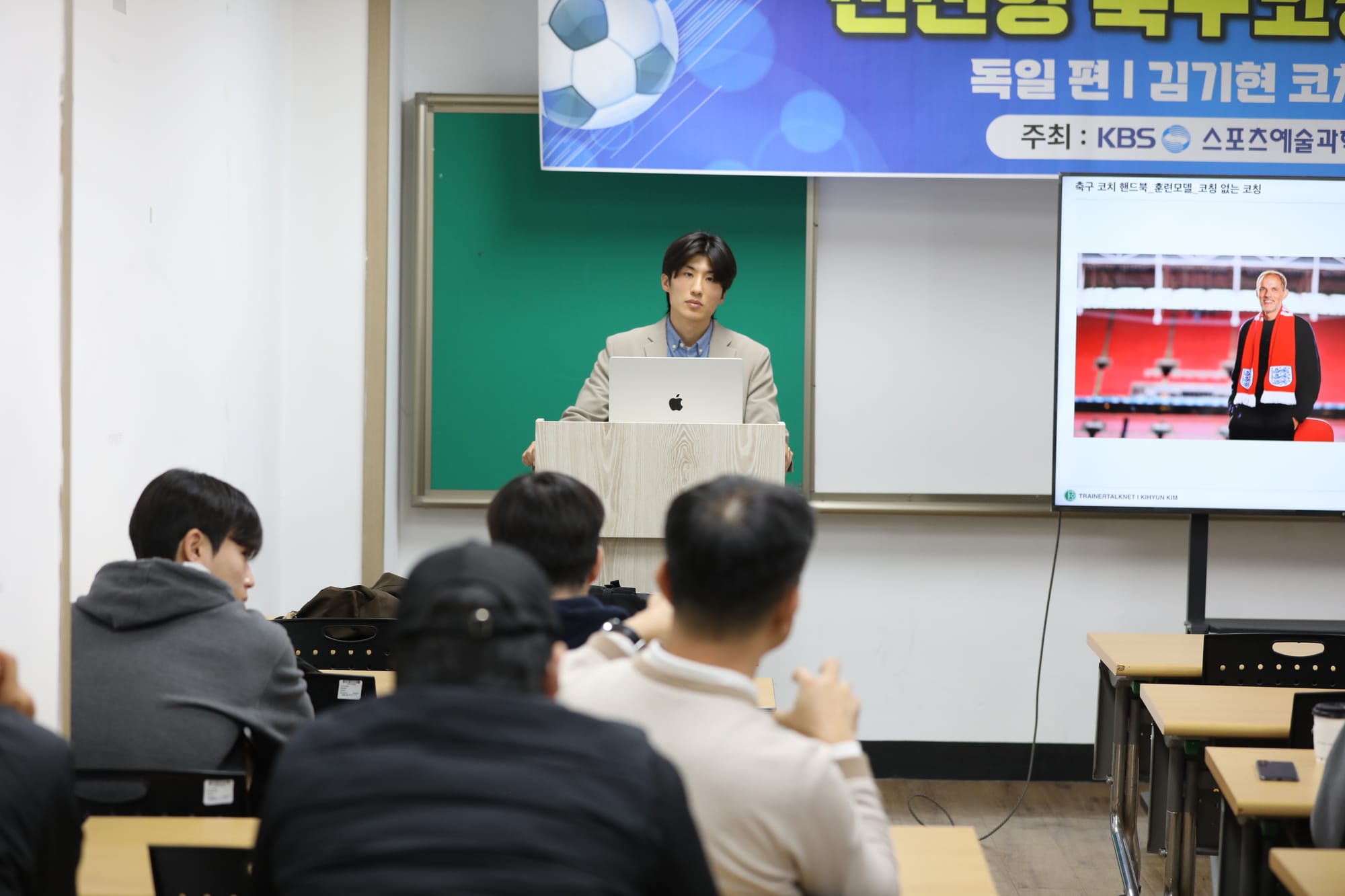 Ki-hyun Kim in a beige blazer speaking at a lectern with a laptop, in front of a lecture banner and a screen, addressing a seated audience.