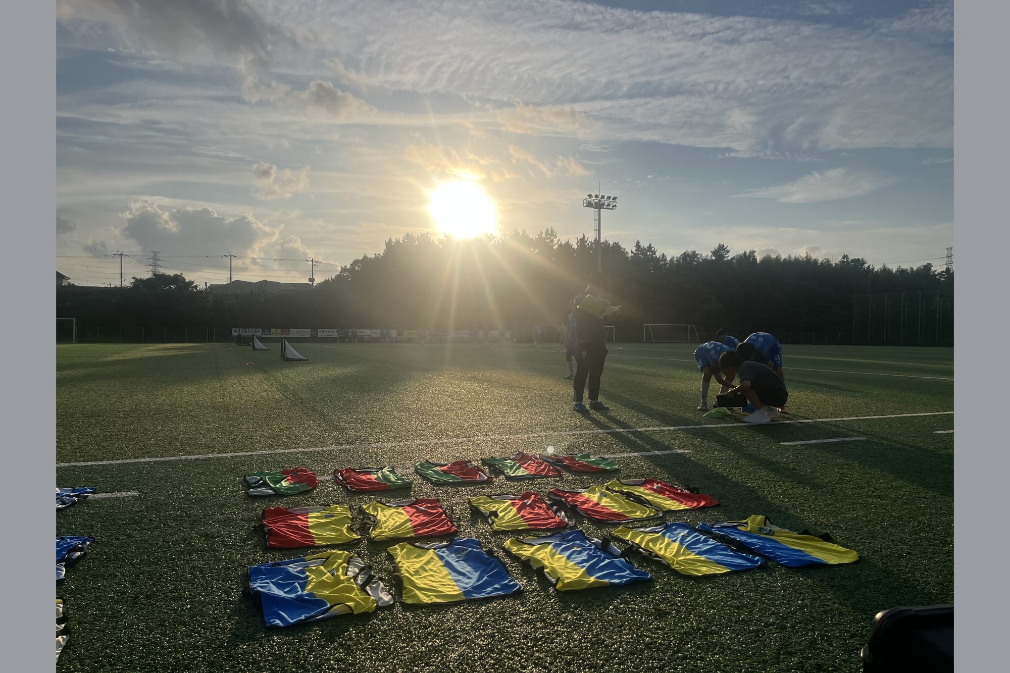 Training bibs in red, yellow, green and blue laid out on a grass pitch at sunset, with coaches and players in the background.
