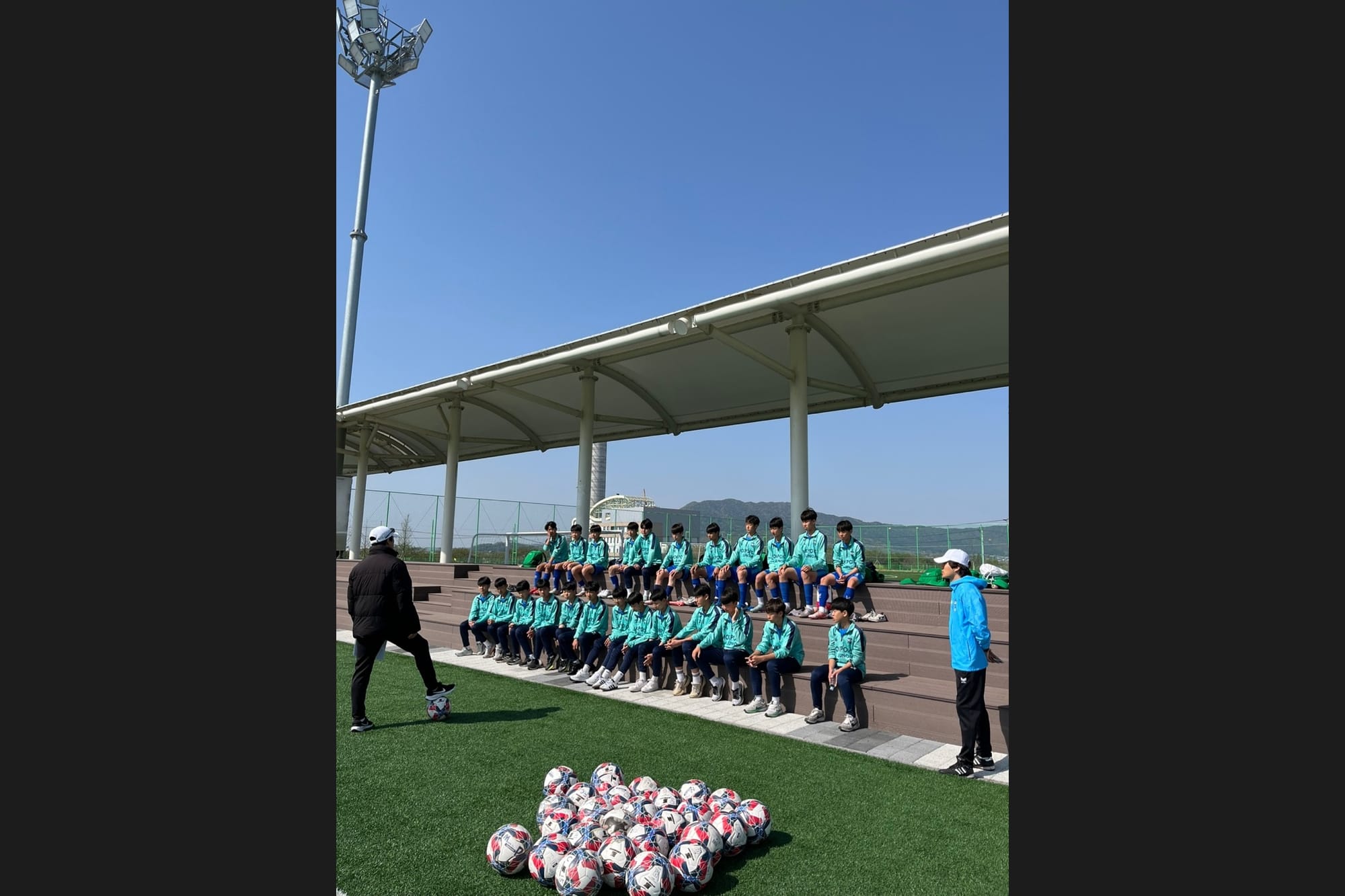 A youth football squad in teal kit seated on covered bleachers at an outdoor pitch, with a coach and a pile of match balls in the foreground.