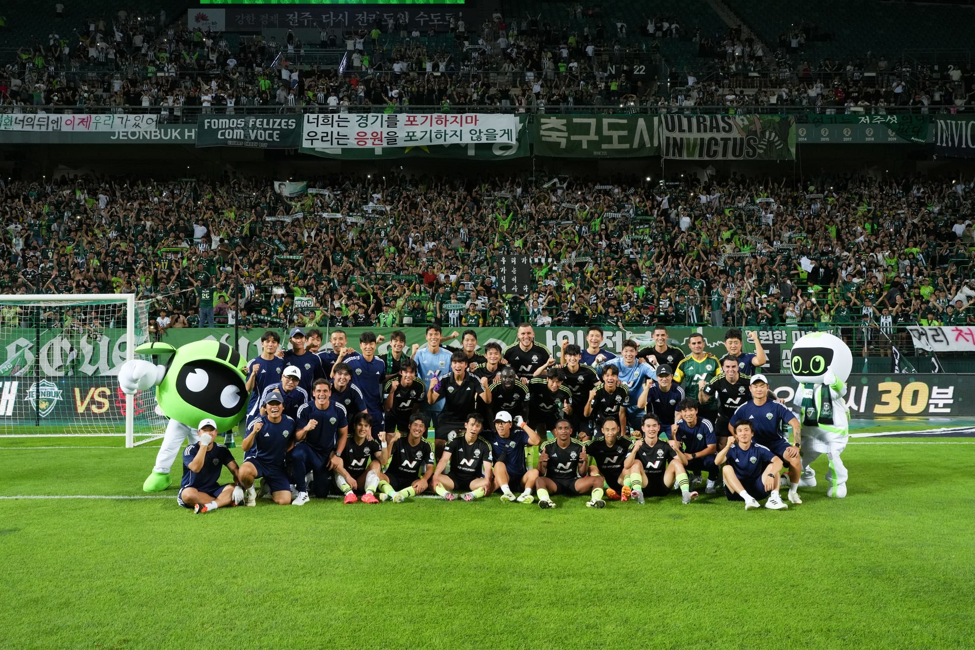 A K League team and staff posing for a group photo on the pitch in front of a packed, banner-covered stand at night.