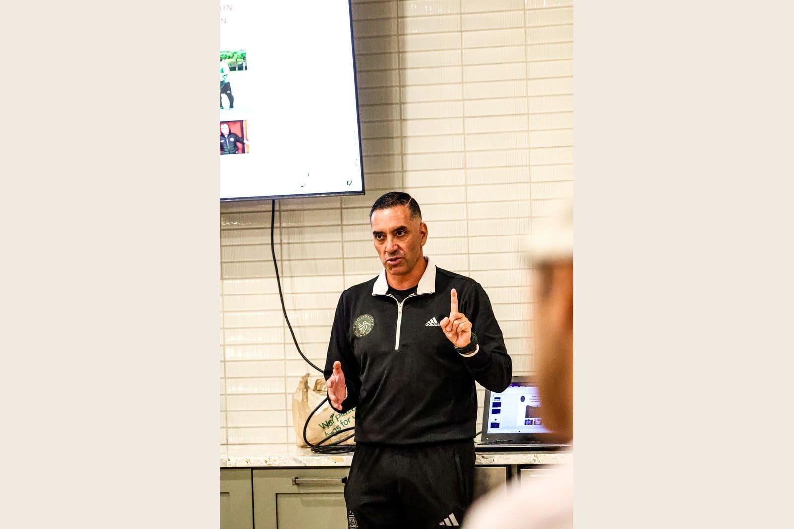 Reinerio Márquez in a black Fort Myers City FC training top with adidas branding, one finger raised, teaching a tactical concept in front of a screen showing coaching footage.