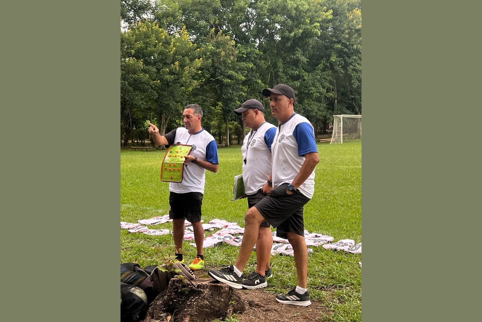 Reinerio Márquez in a blue-and-white training top holding a tactical board with red and yellow figurines, briefing two other staff members outdoors on a grass pitch surrounded by trees.