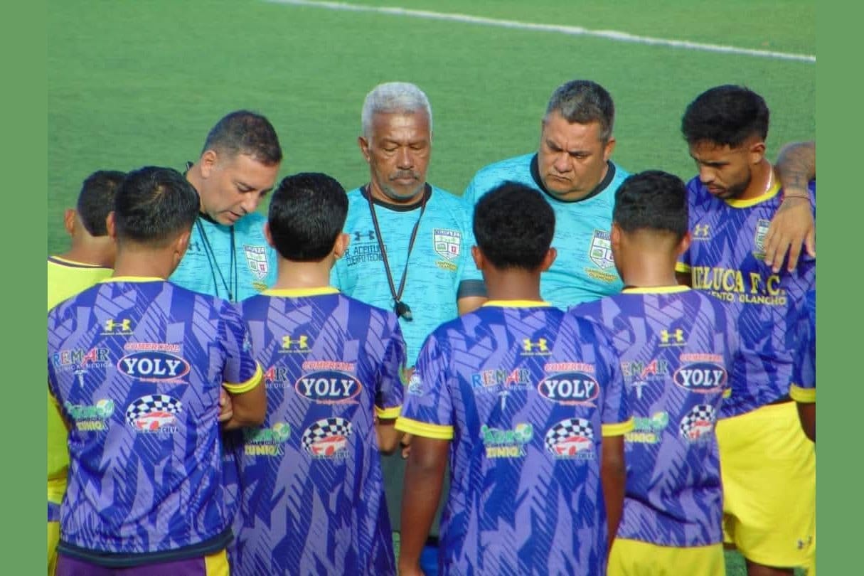 Reinerio Márquez, in a light blue Meluca FC coaching shirt, standing in a huddle with young Meluca players in purple-and-yellow kits, all with heads bowed for a pre-match team moment.