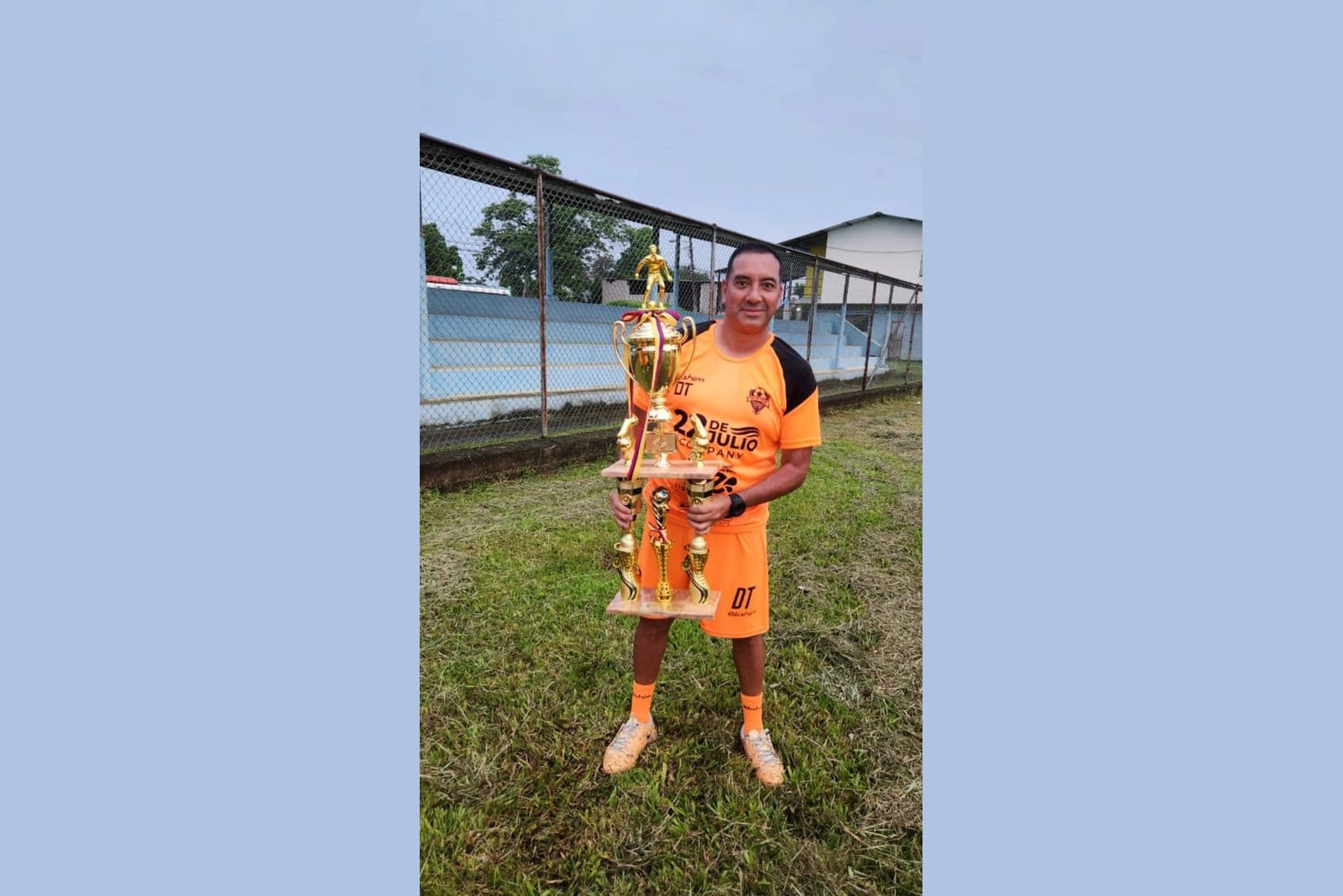 Reinerio Márquez, in an orange 22 de Julio FC coaching kit with "DT" (Director Técnico) on the chest, holding a tall gold trophy in front of a stadium fence after winning the Tumaco International Invitational.