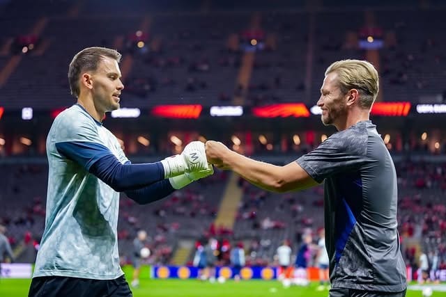 Jyri Nieminen fist-bumping a Feyenoord goalkeeper wearing gloves before a match, with the empty stadium stands visible in the background.