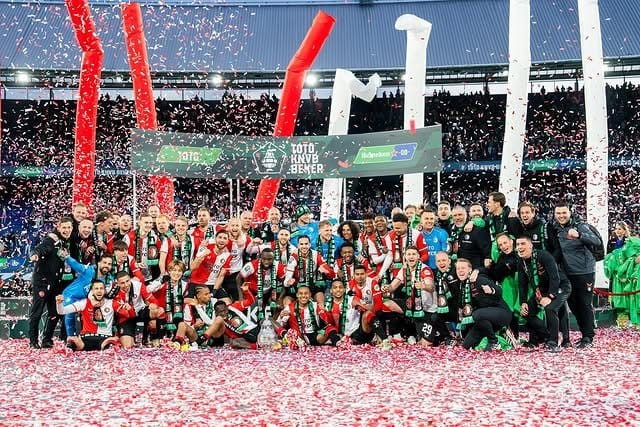 Feyenoord players and coaching staff posing for a KNVB Beker championship photo under falling red-and-white confetti, with the trophy at the centre of the group and Jyri Nieminen visible among the coaching staff on the right.