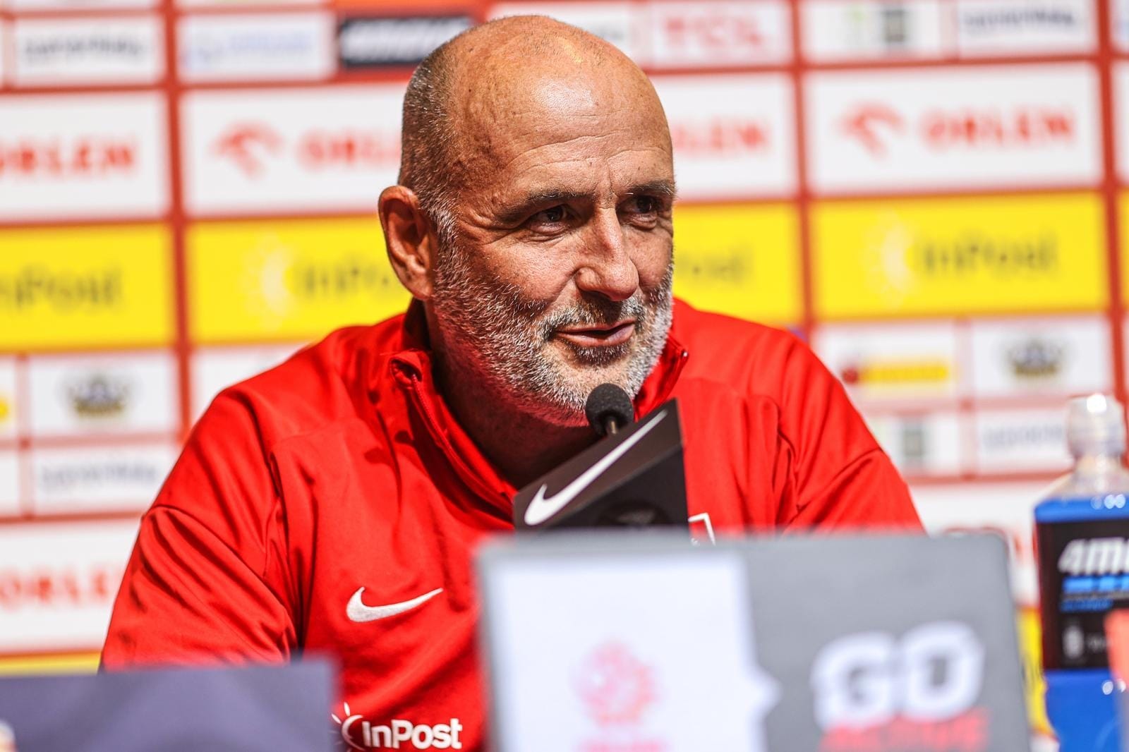 Michał Probierz in a red Poland training top, speaking at a press conference behind a microphone with sponsor boards in the background.