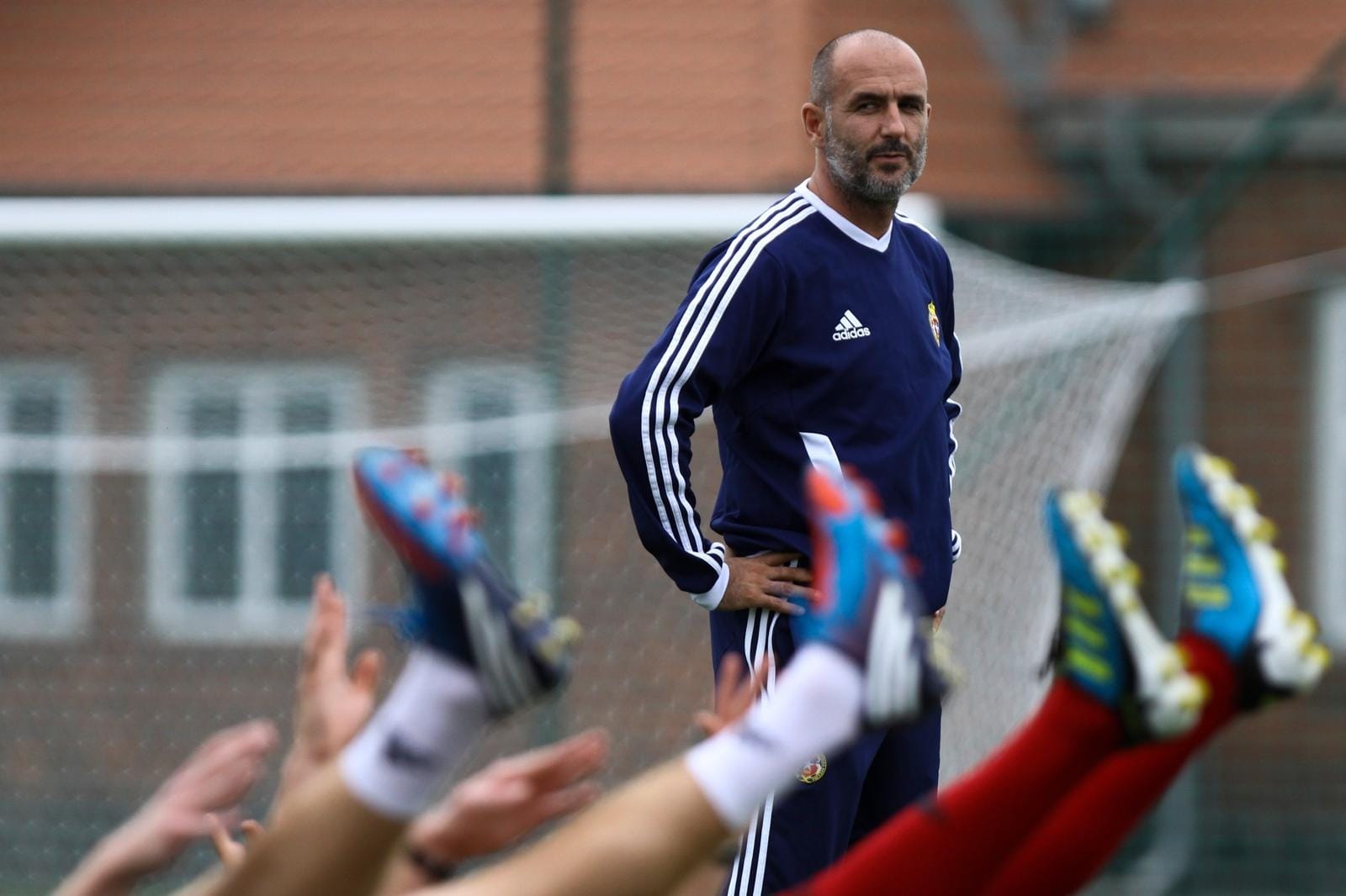 Michał Probierz in a navy adidas training top, hands on his hips, watching players perform stretching exercises on a training pitch.