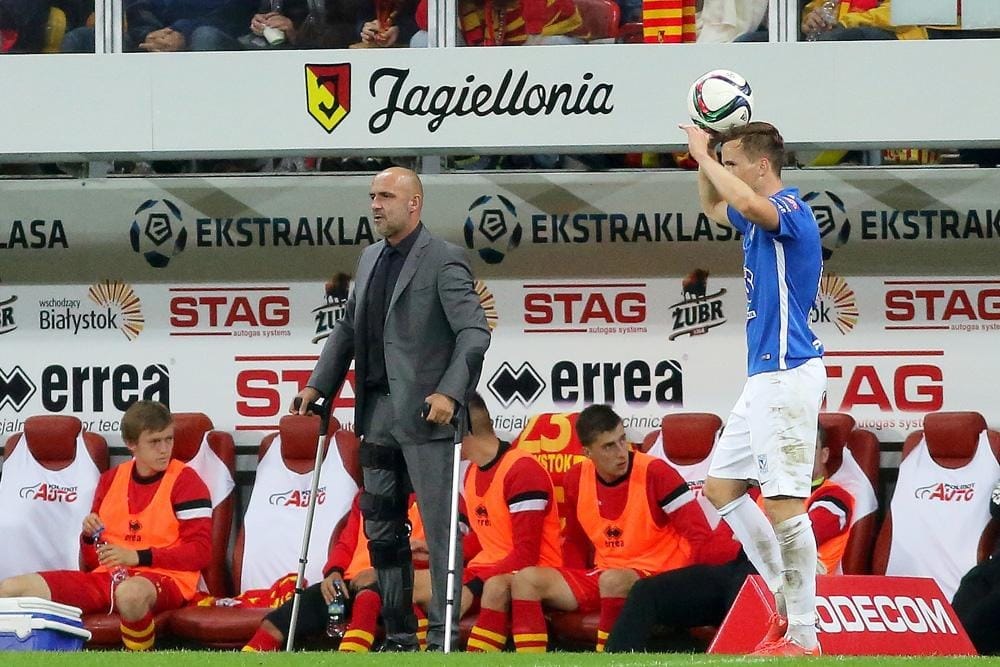Michał Probierz in a grey suit, standing on crutches at the edge of the technical area during an Ekstraklasa match as a player prepares a throw-in.