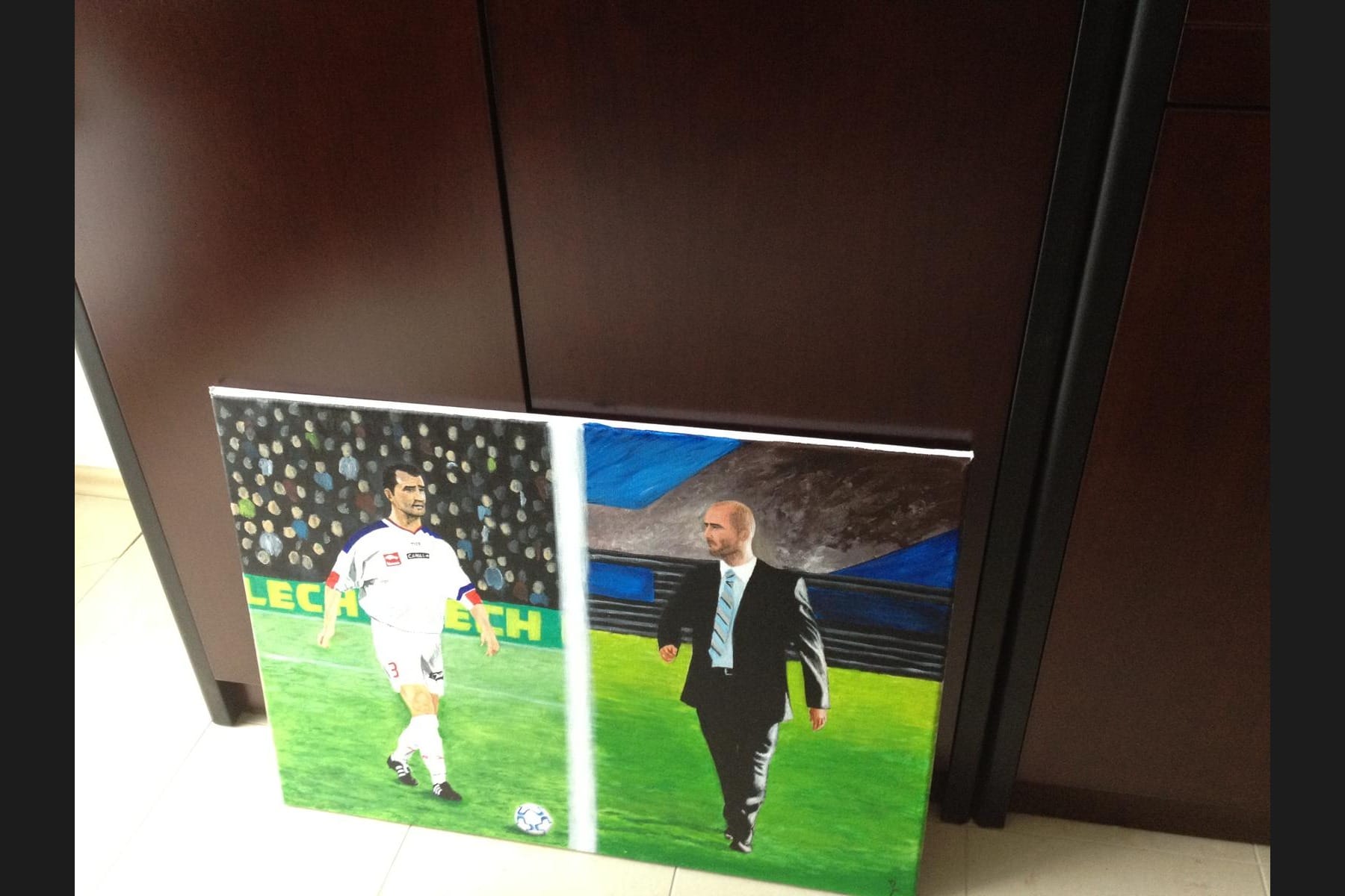 A painted canvas showing Michał Probierz twice — as a footballer in a white kit and as a coach in a suit walking the pitch — leaning against dark furniture.