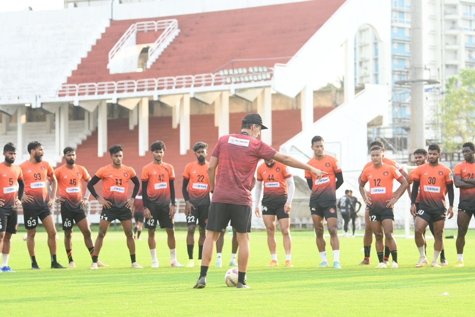 Domingo Oramas, seen from behind in a cap and training kit, addressing a line of players in orange during a session at a stadium.