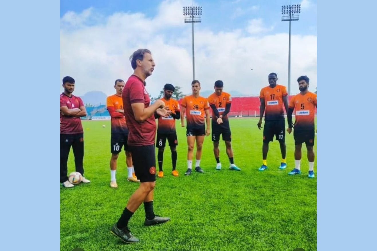 Domingo Oramas in a dark-red training top speaking to a group of players in orange kit on a grass pitch.