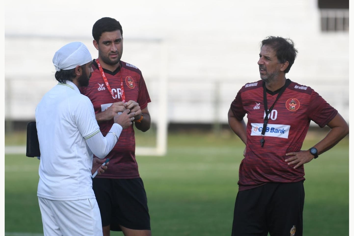 Domingo Oramas in a dark-red training top talking with a staff member and a player at the side of a pitch.