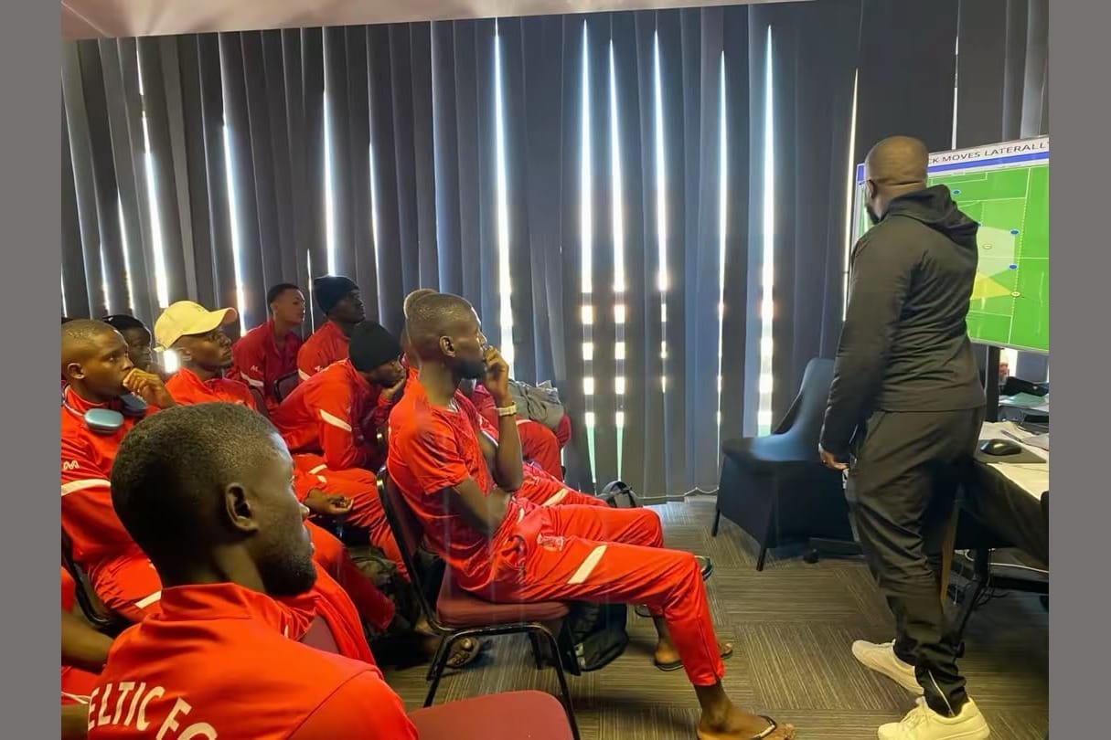 Mark Jackson, in a dark tracksuit, presenting a tactical diagram on a screen to his seated Celtic FC squad in red training kit.