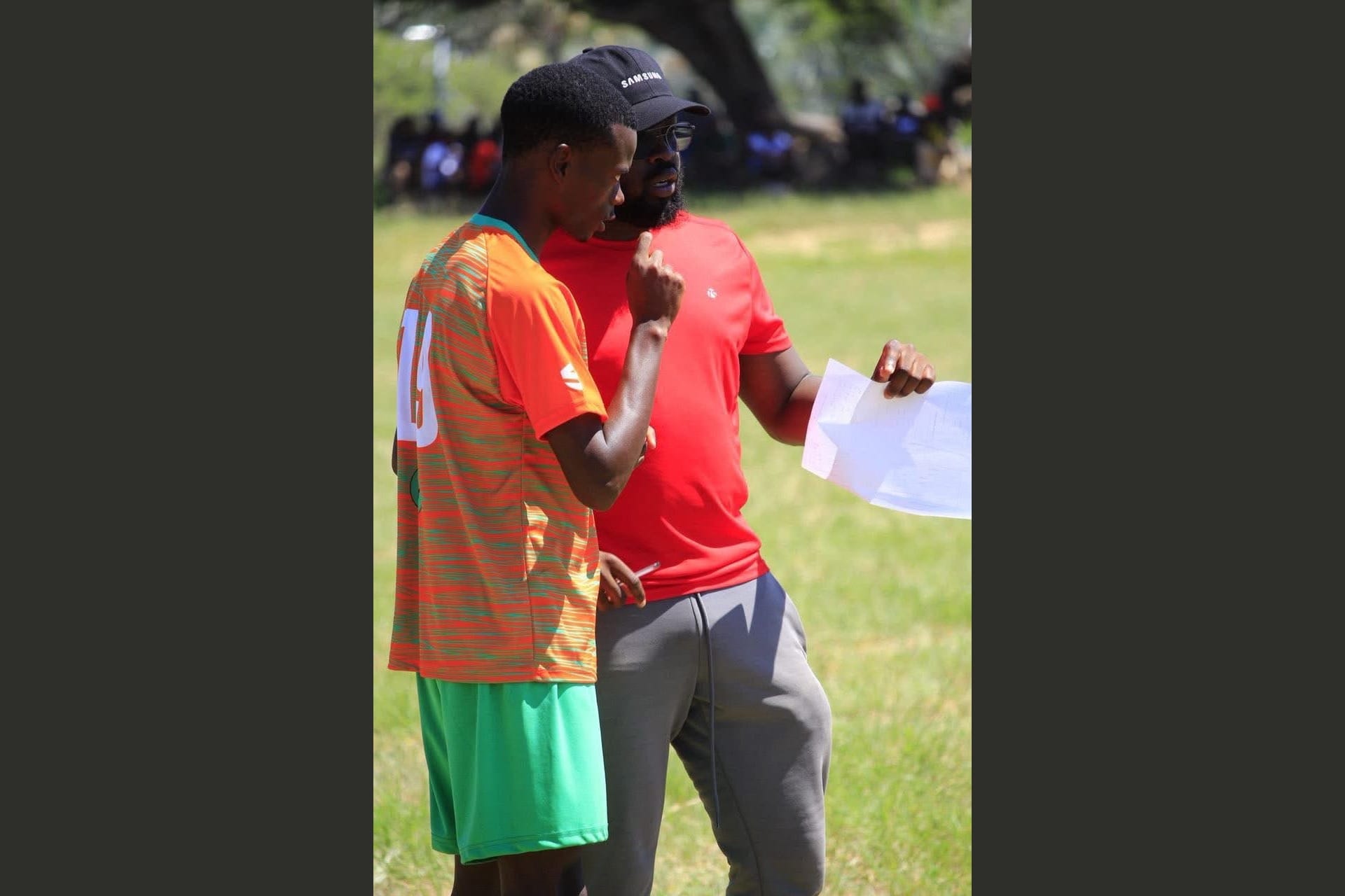Mark Jackson in a red shirt and cap talking closely with a young player in an orange kit, holding notes, on a grass pitch.