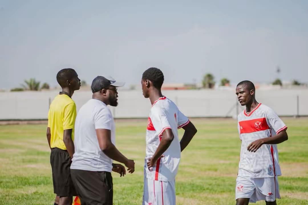 Mark Jackson in a white shirt and cap speaking with players in red-and-white kit and a referee during a match on a grass pitch.