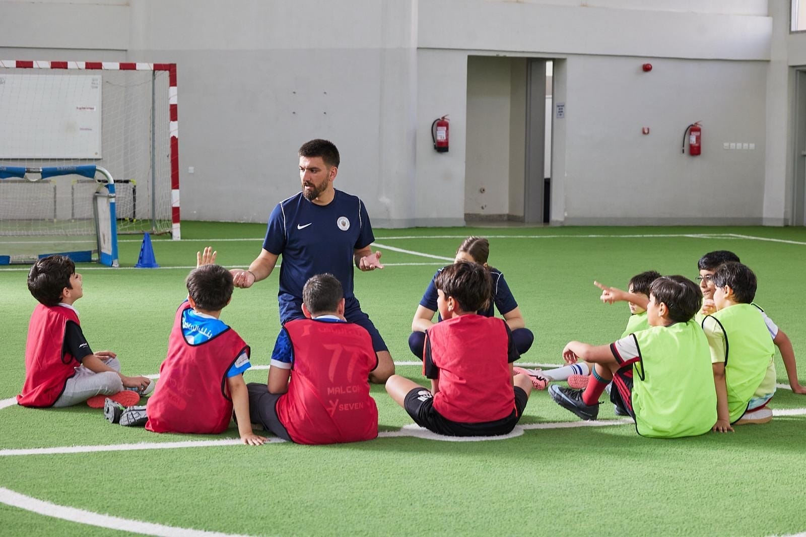 Simone Canepa crouching to speak with a circle of young children in coloured training bibs on an indoor pitch.