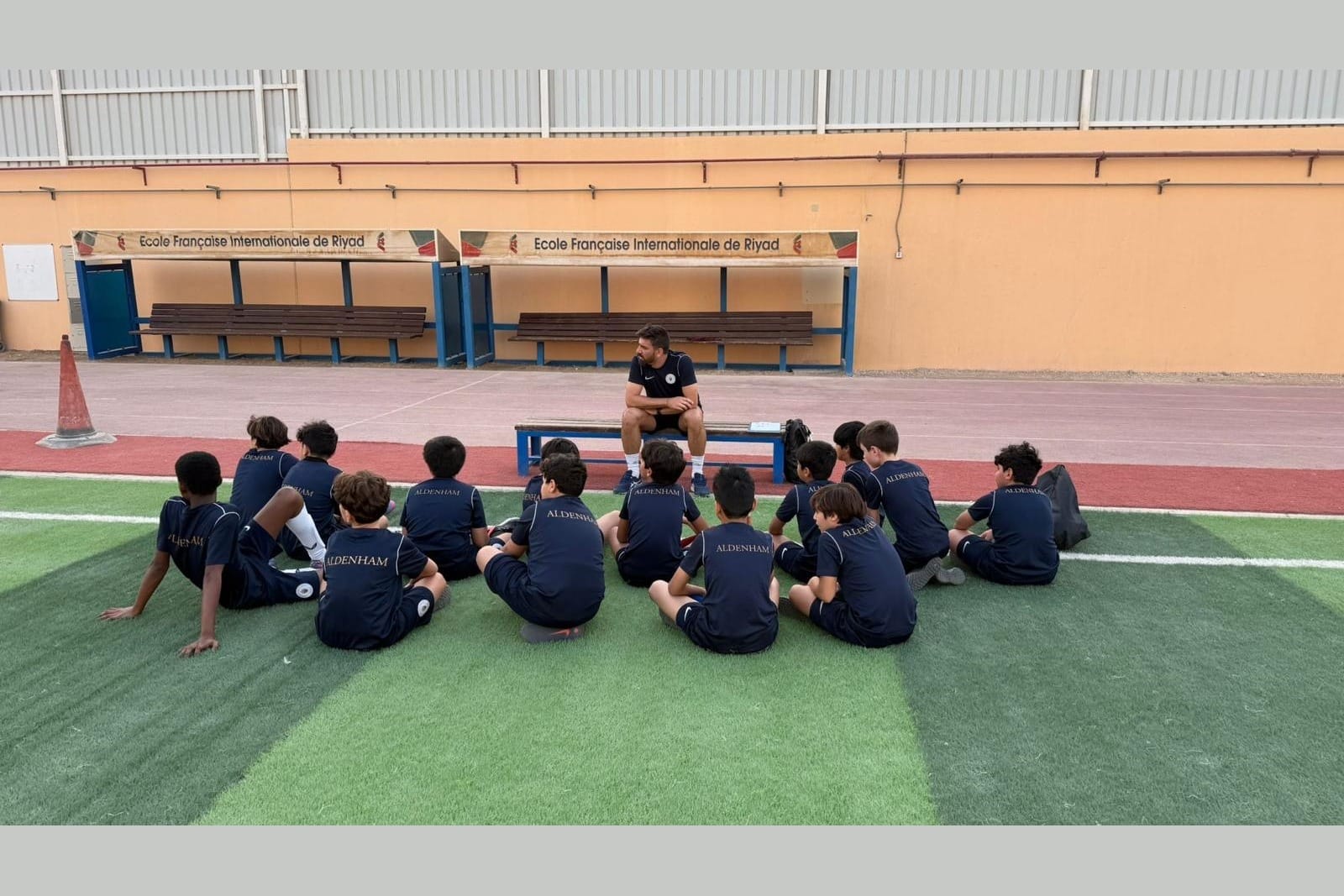 Simone Canepa seated on a bench addressing a group of children in navy Aldenham kit seated on the grass around him.