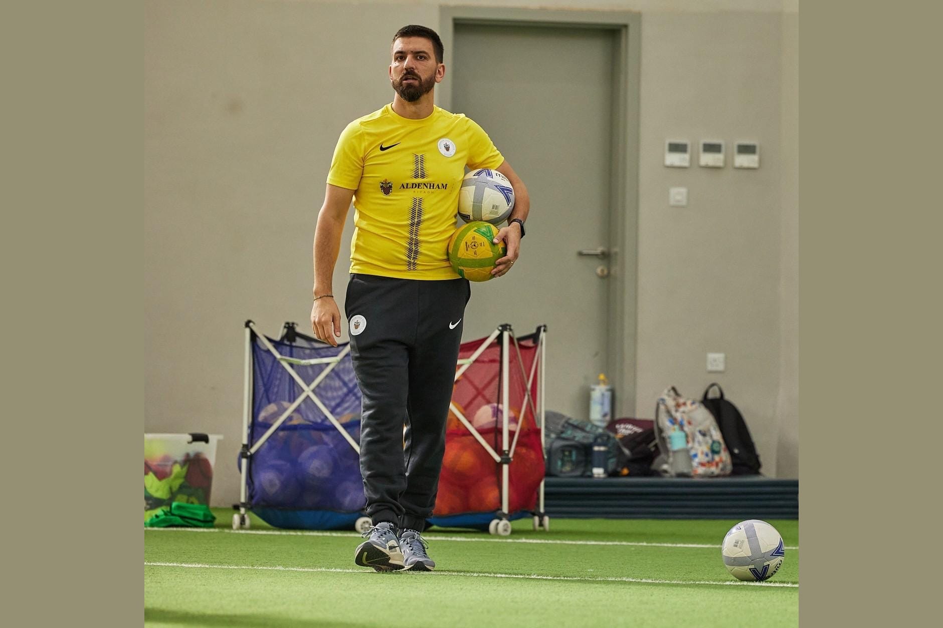 Simone Canepa in a yellow Aldenham training top holding two footballs on an indoor pitch, with ball baskets behind him.