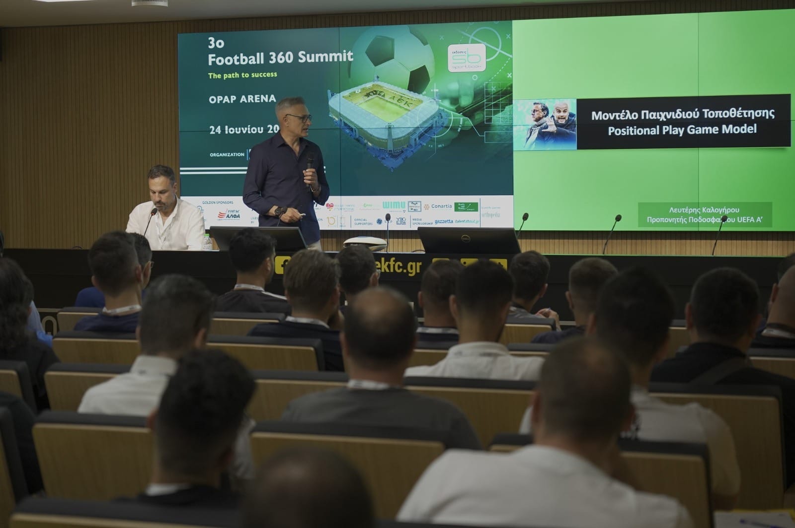 Lefteris Kalogirou speaking into a microphone at a football conference, in front of a large screen reading "Positional Play Game Model".