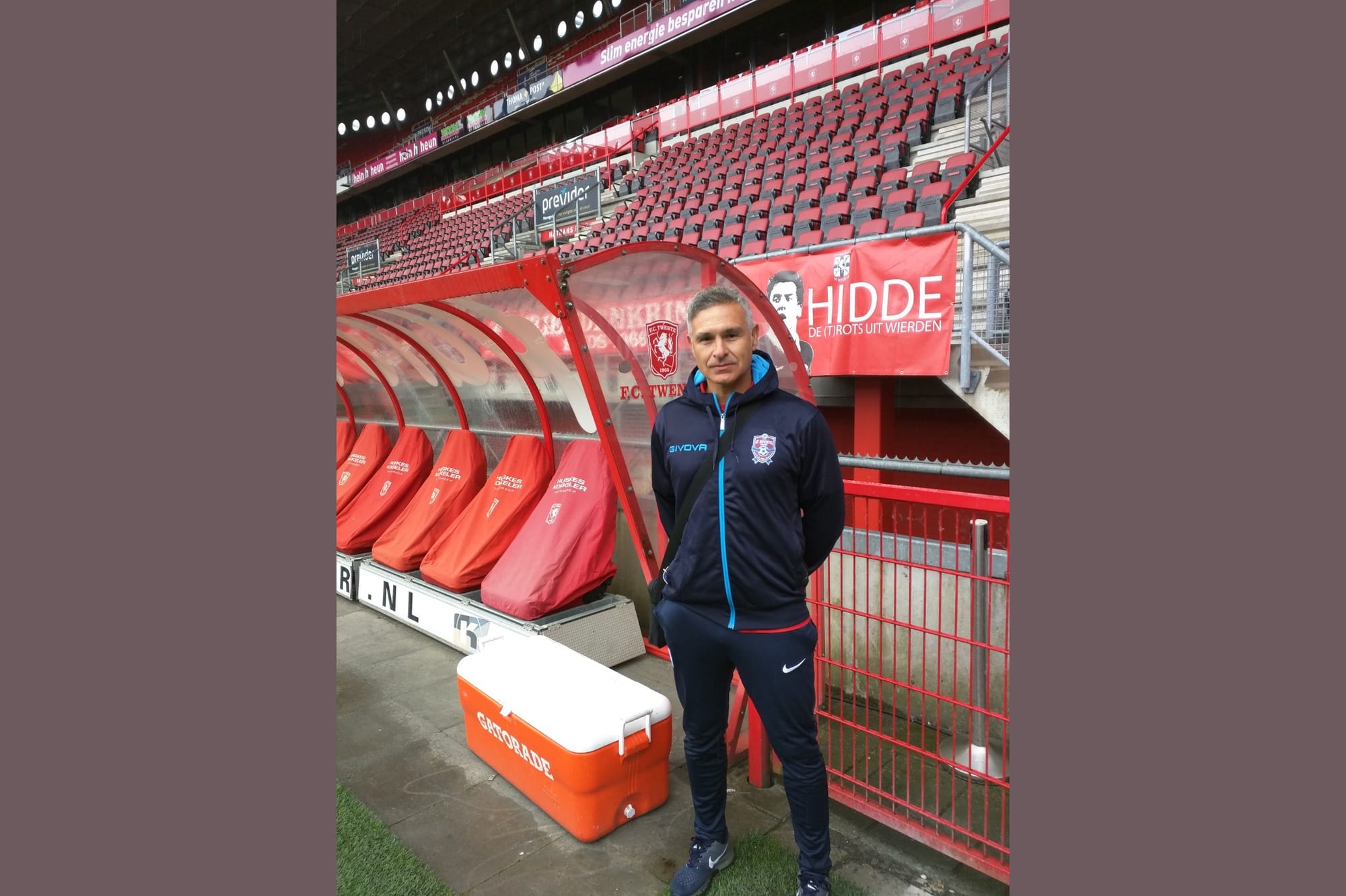 Lefteris Kalogirou standing beside the covered dugout inside FC Twente's stadium in a navy tracksuit.