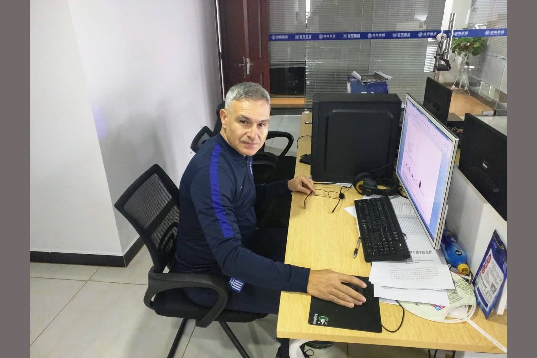 Lefteris Kalogirou seated at a desk with a computer in an office in China, turning toward the camera in a navy tracksuit.