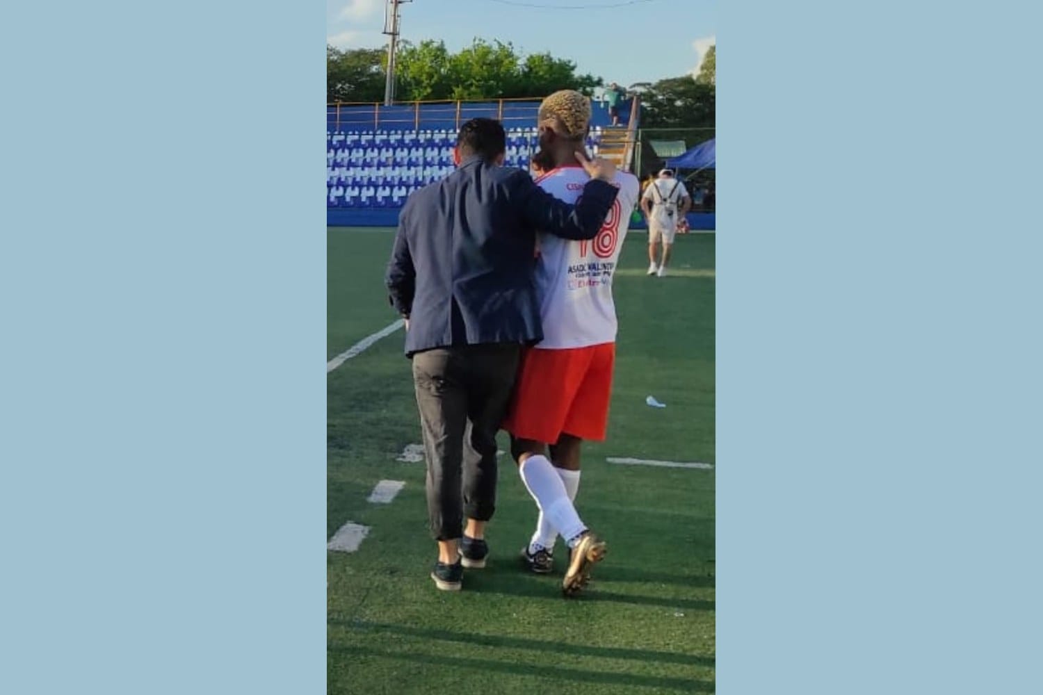 Manuel Sánchez in a navy jacket, seen from behind with his arm around a Deportivo Masaya player wearing a white and red jersey with the number 18, walking off the pitch together.