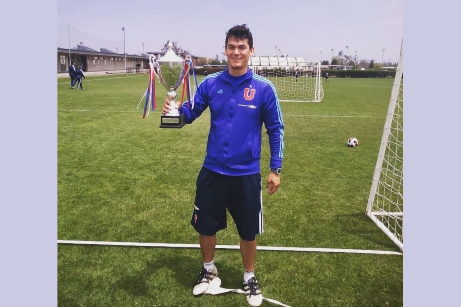 A younger Manuel Sánchez in a blue Universidad de Chile track jacket holding a small trophy on a training pitch with a white goalpost in the background.