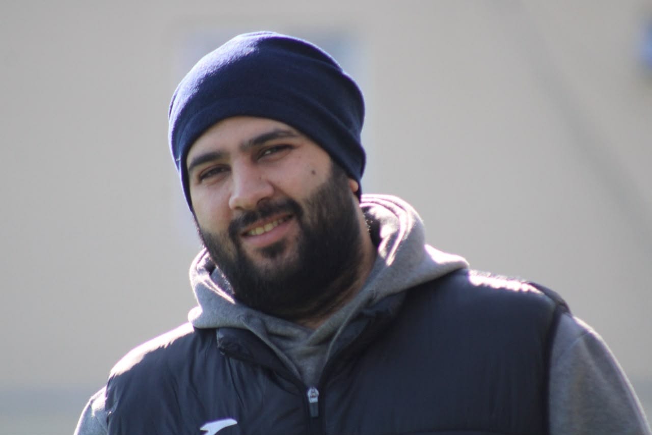 Tahir Salayev in a beanie and dark gilet, smiling outdoors on a bright day at a training ground.