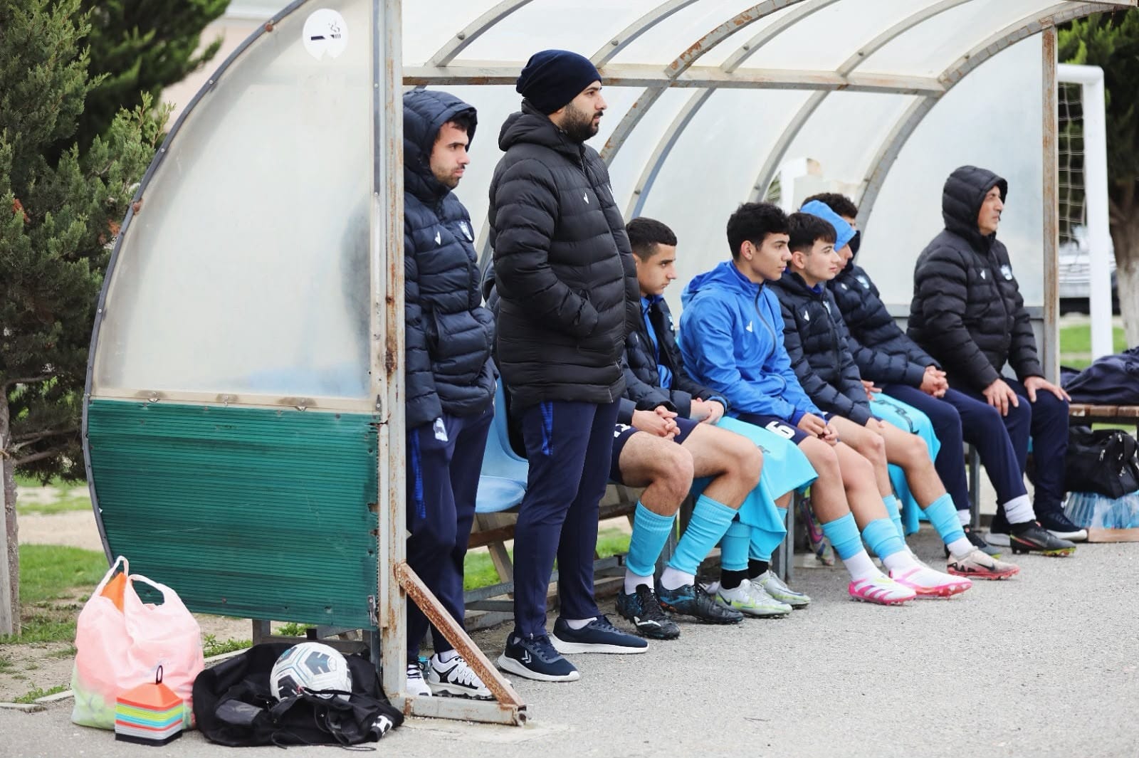 Tahir Salayev in a black coat and beanie standing at the front of a dugout as young players in blue kit sit watching the match.