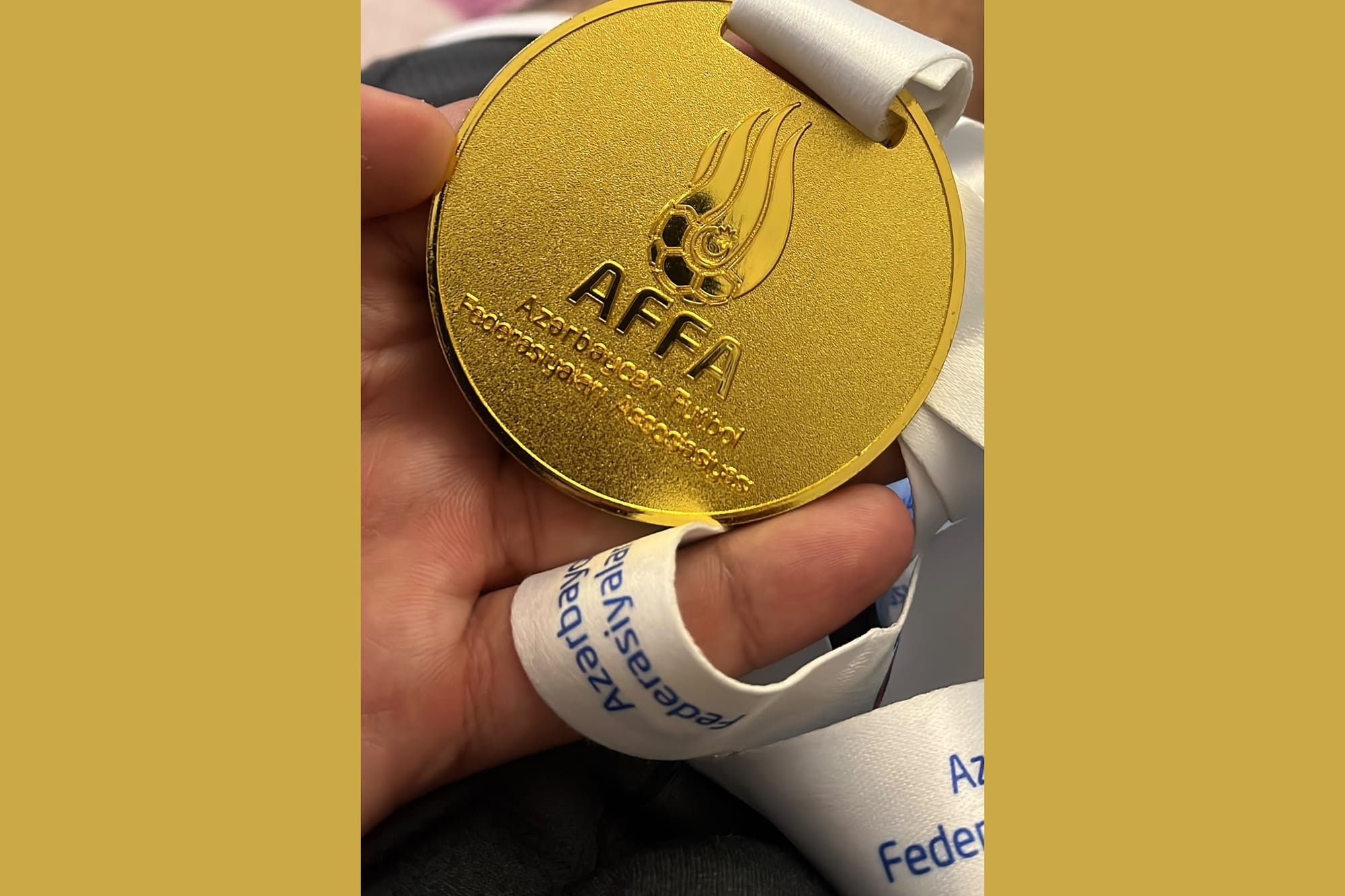 A close-up of a gold AFFA (Azerbaijan Football Federation) winners' medal on a white ribbon, held in one hand.