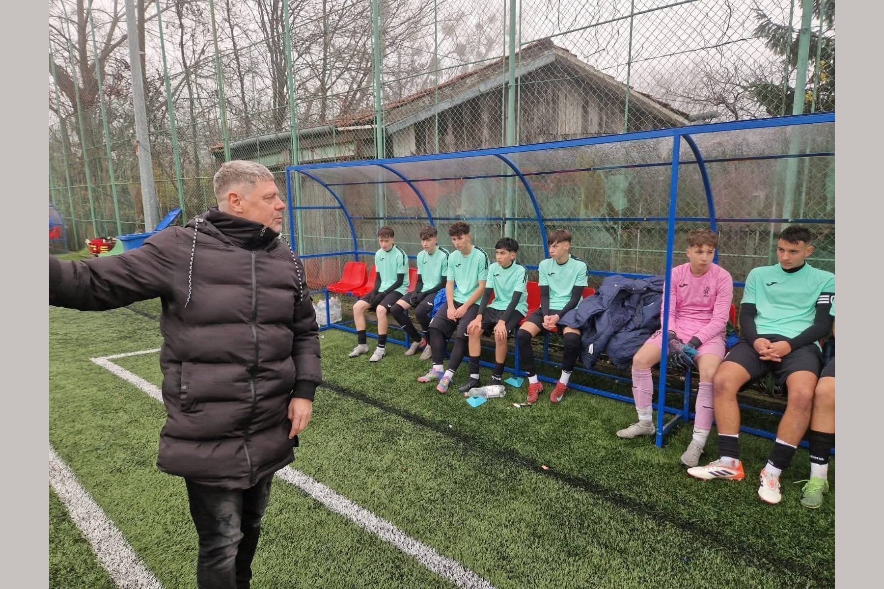 Tibor Selymes in a black coat gesturing to a row of young players in green kit seated in a dugout at a training ground.