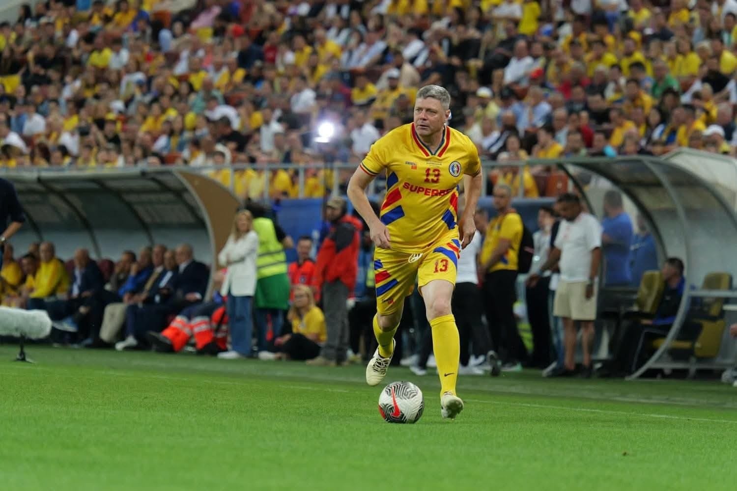 Tibor Selymes in Romania's yellow number 13 shirt running with the ball during a legends match in a packed stadium.